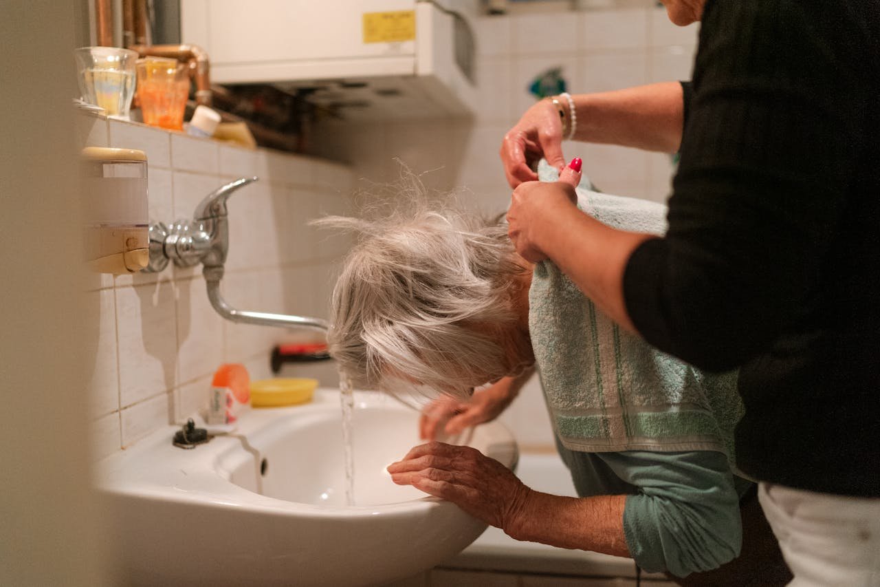 Elderly woman with gray hair receiving help from a caregiver in a bathroom setting.